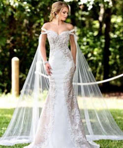 Woman in an off-the-shoulder, lace wedding gown with long, sheer cape stands outdoors on grass.