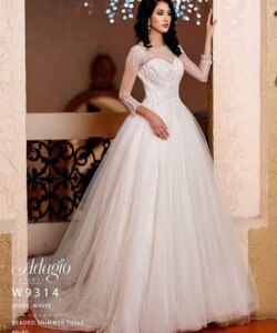 Woman wearing an ivory white beaded shimmer tulle ball gown with sheer sleeves, standing indoors by an archway.