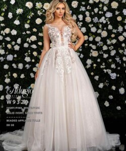 A woman in a white, beaded tulle ball gown stands in front of a wall covered with white and green flowers.