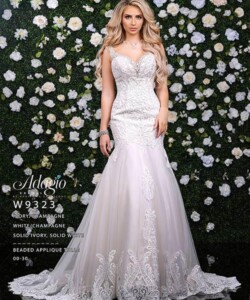 Woman in a beaded ivory wedding gown stands in front of a flower-covered wall, looking directly at the camera.