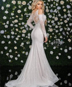 A woman in a long, white, lace gown with off-the-shoulder sleeves stands in front of a flower-covered wall.