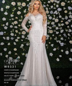 A woman in a fitted, off-shoulder, embroidered ivory gown stands in front of a flower-covered wall.