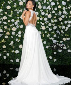 Woman in a white, backless gown stands in front of a wall covered with white roses and greenery.