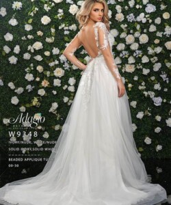 Woman in an open-back, long-sleeve white bridal gown stands before a flower-covered wall, facing sideways.