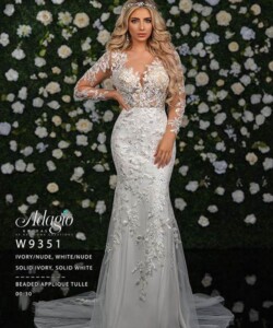 Woman wearing a long-sleeve, lace-embellished white wedding gown stands in front of a floral wall.