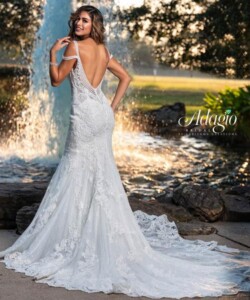 Woman in a fitted, lace, off-shoulder wedding dress with a long train, standing by a fountain outdoors.