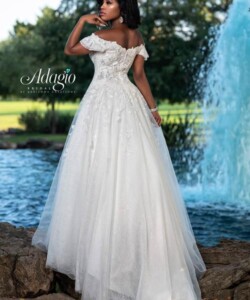 A woman in an off-shoulder white bridal gown stands by a pond and fountain, facing away from the camera.