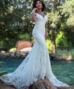 A woman in a fitted, white lace wedding dress stands outdoors near water and greenery.