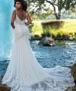 A woman in a lace wedding gown stands outdoors near a pond and fountain, facing away from the camera.