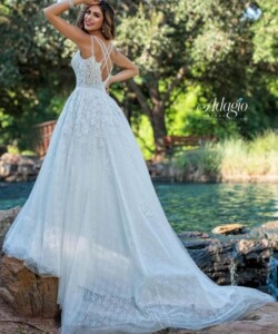 A woman in a white lace wedding dress stands on rocks near a pond with trees in the background.