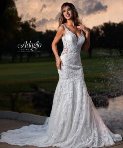 A woman wearing a white lace wedding dress stands outdoors near a pond at sunset.