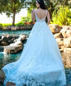 A woman in a white lace wedding gown stands by a pond with rocks and greenery in the background.