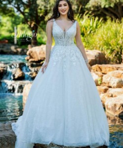 A woman in a white, embellished bridal gown stands by a small waterfall and rocks in a garden setting.