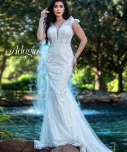 Woman in a white, lace wedding dress poses outdoors in front of a water fountain and green trees.