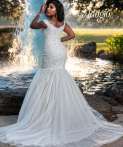 A woman in a fitted white lace wedding gown poses outdoors near a rock-lined fountain and greenery.
