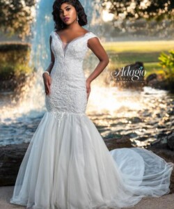 Woman in a fitted white lace mermaid wedding gown stands outdoors near a fountain, with greenery in the background.