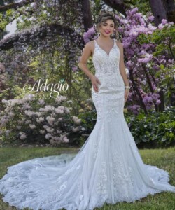 A woman in a white lace wedding dress stands on grass in front of blooming pink and white flowers.