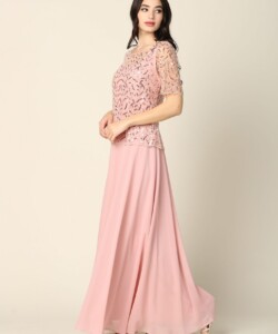 Woman wearing a light pink, floor-length dress with a patterned lace top and short sleeves, standing against a plain background.