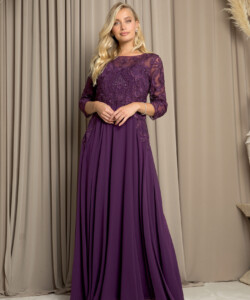 Woman wearing a long-sleeved, floor-length purple gown with lace details, standing in front of beige curtains.