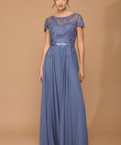 A woman wearing a floor-length blue gown with lace detailing on the bodice and short sleeves stands against a tan background.